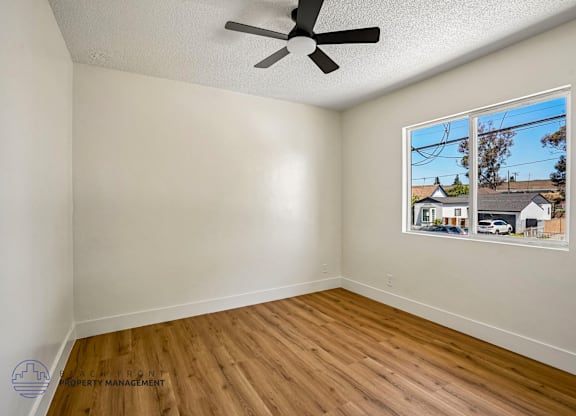 A room with a ceiling fan and a window showing a view of houses.
