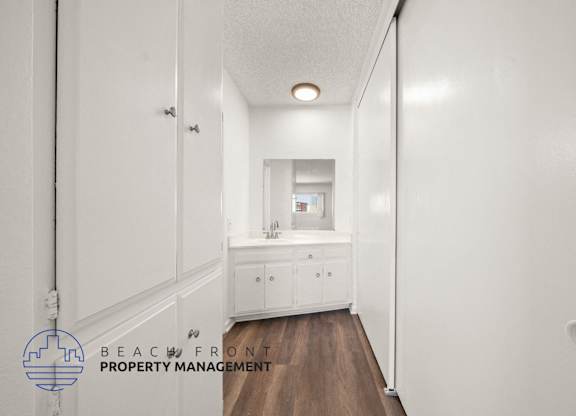 A hallway with white walls and a wooden floor leads to a bathroom.