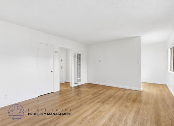 A spacious room with wooden flooring and white walls, with a door on the left and a window on the right.