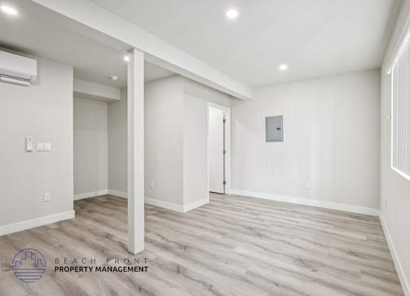A spacious room with wooden flooring and white walls, with a logo for Beach Front Property Management in the bottom left corner.