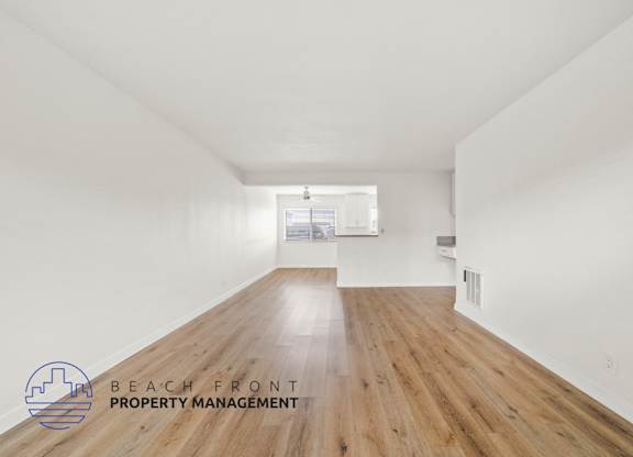 A spacious room with wooden floors and white walls, with a logo for Beach Front Property Management in the bottom left corner.