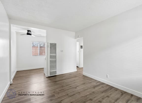 A spacious room with wooden floors and white walls, featuring a ceiling fan and a window with blinds.