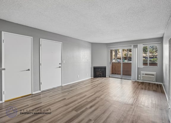 A spacious room with wooden floors and white walls, featuring a fireplace and a window with a view of trees.