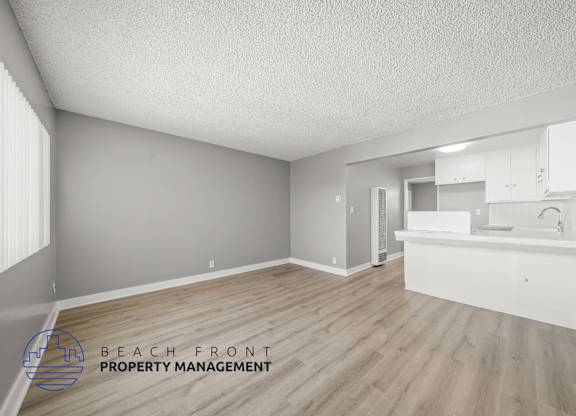 A spacious room with wooden flooring and a kitchen area with white cabinets.