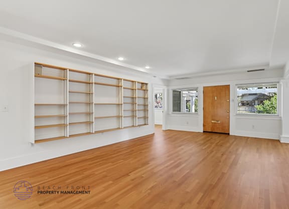 A spacious room with wooden floors and white walls, featuring a large window with blinds and a wooden door.