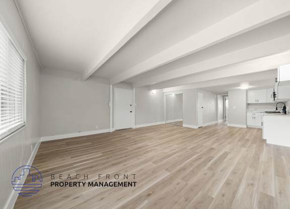 A spacious room with wooden flooring and white walls, with a logo for Beach Front Property Management in the bottom left corner.