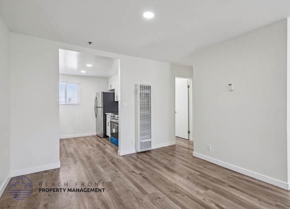 A spacious room with wooden flooring and white walls, featuring a kitchen area with a refrigerator and a sink.