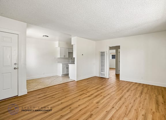 A spacious room with wooden floors and white walls, with a door on the left and a kitchen area visible in the distance.