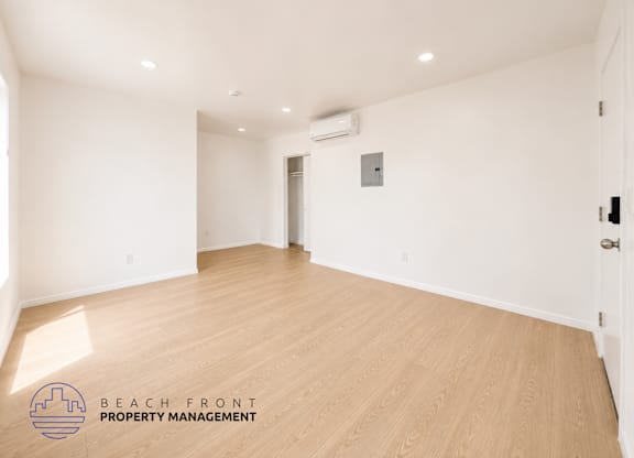 A spacious room with wooden flooring and white walls, with a logo for Beach Front Property Management in the bottom left corner.