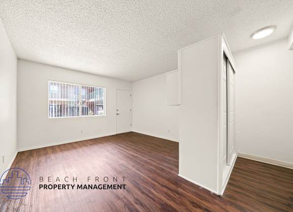 A room with wooden floors and white walls, with a window showing a view of a building.
