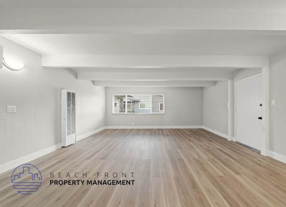 A spacious room with wooden flooring and white walls, with a logo for Beach Front Property Management in the bottom left corner.