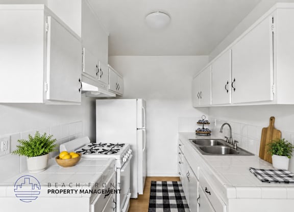 A kitchen with white cabinets and appliances, a black and white checkered mat on the floor, and a logo for Beach Front Property Management in the corner.