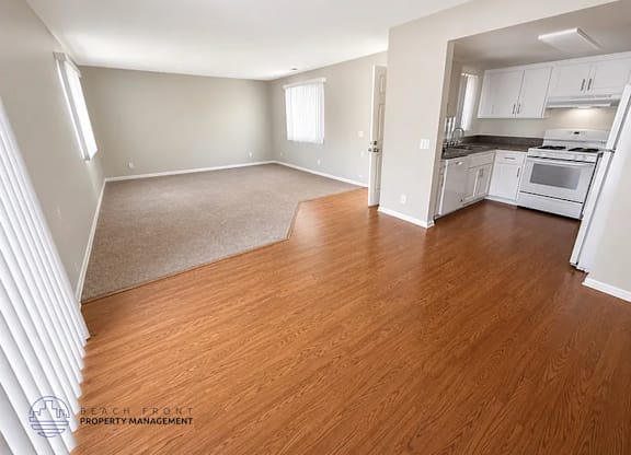 A spacious living room with wooden floors and a kitchen area in the background.