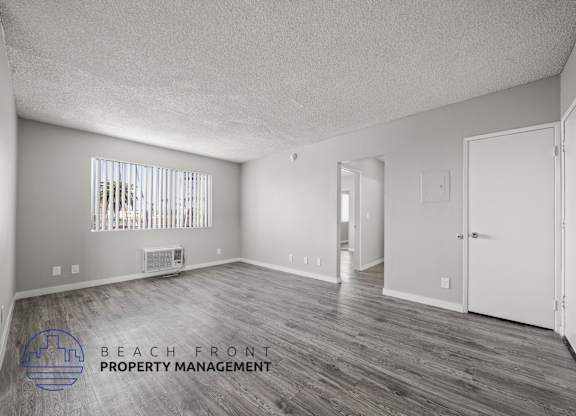 A spacious room with wooden flooring and white walls, with a window covered by vertical blinds.