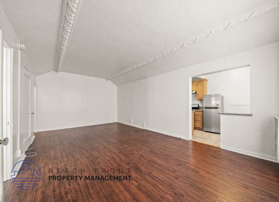A spacious room with wooden floors and white walls, with a kitchenette visible in the background.