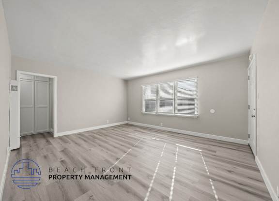 A spacious room with wooden flooring and a window with blinds.