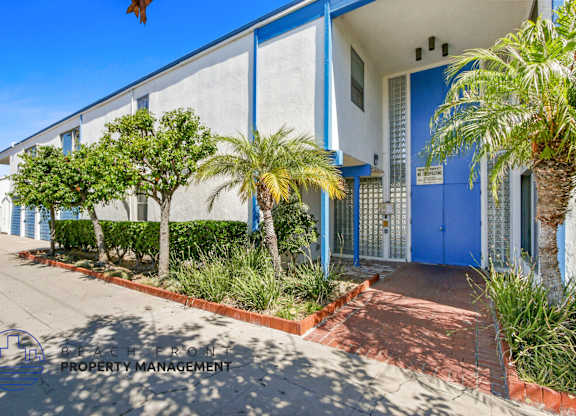 A blue building with a white door and windows, surrounded by greenery and a red brick pathway.