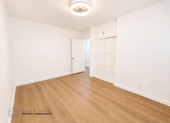A spacious room with wooden flooring and white walls, with a logo for Beach Front Property Management in the bottom left corner.