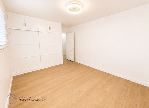A white room with wooden flooring and a logo for Beach Front Property Management in the bottom left corner.