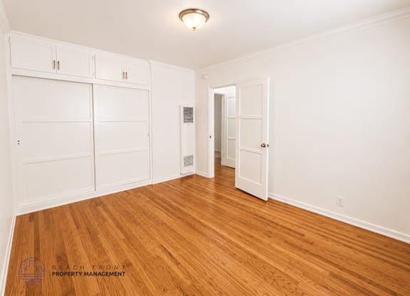 A spacious room with wooden floors and white walls, featuring a set of white built-in cabinets and a doorway leading to another room.