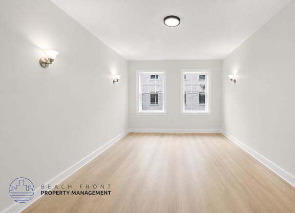 A spacious room with wooden floors and white walls, featuring a large window with a view of a building outside.