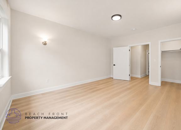 A spacious room with wooden flooring and white walls, with a logo for Beach Front Property Management in the bottom left corner.