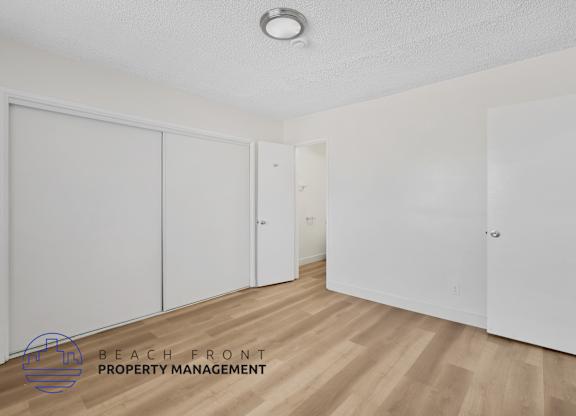 A room with white walls and wooden flooring, with a logo for Beach Front Property Management in the bottom left corner.