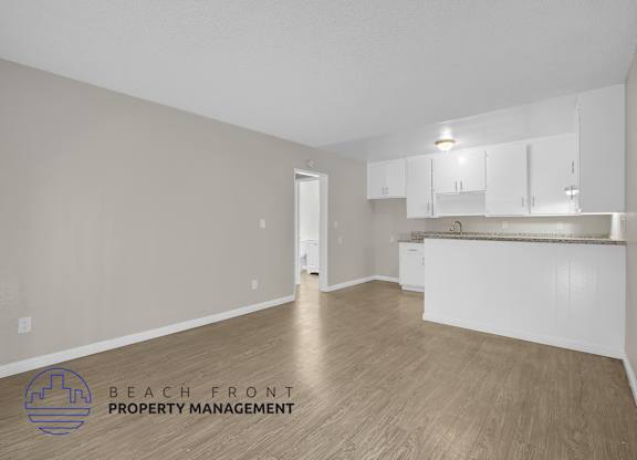 A spacious room with wooden flooring and white walls, featuring a kitchen area with white cabinets and a countertop.