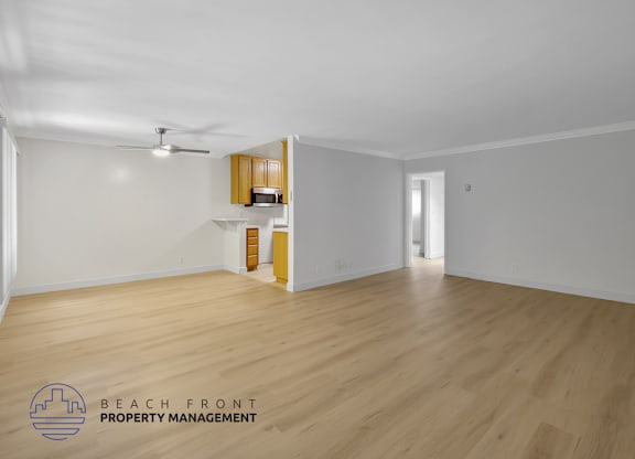 A spacious room with wooden flooring and white walls, featuring a kitchenette with wooden cabinets and a ceiling fan.