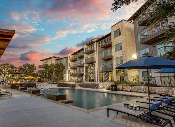 A swimming pool surrounded by lounge chairs and umbrellas in front of a modern apartment building.