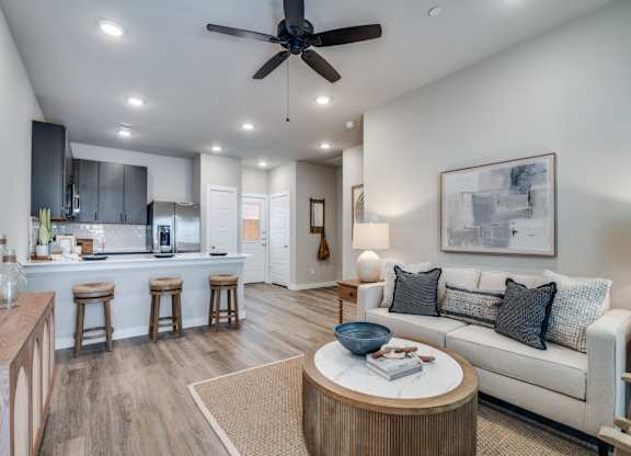 A modern living room with a ceiling fan and a kitchen area in the background.