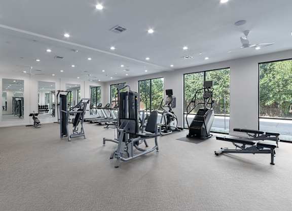 A spacious gym with a variety of equipment and a view of trees outside the windows.