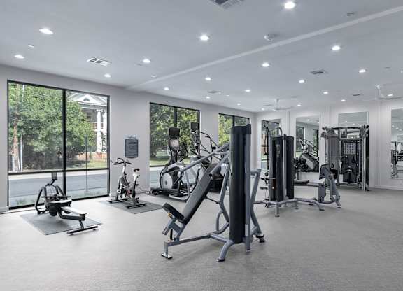 A spacious gym with a variety of equipment and a view of trees outside the windows.