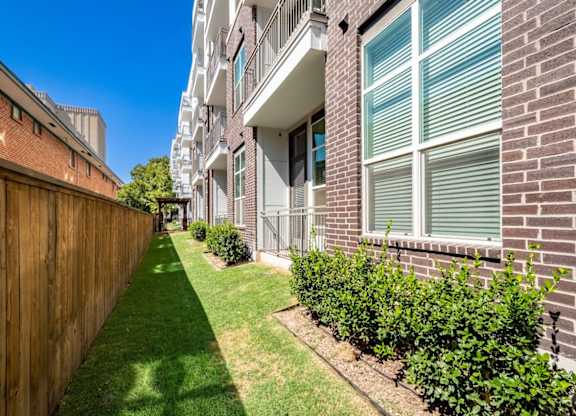 A long, narrow, green lawn separates a brick building from a wooden fence.
