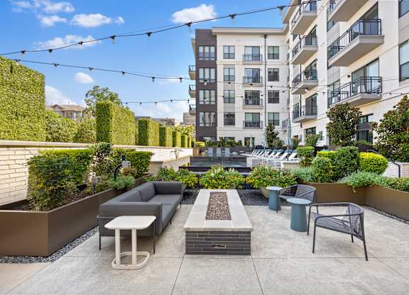 A patio with a table, chairs, and a bench is surrounded by a hedge and a building in the background.