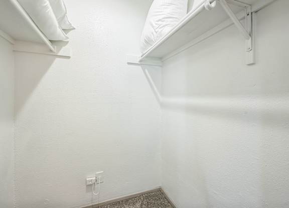 A white wall with a small electrical outlet and a white shelf above a carpeted floor.