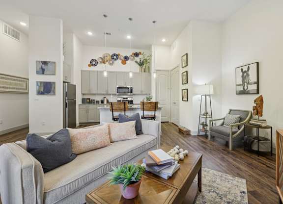An apartment living room featuring a plush beige sofa and warm decor, looking back toward the bright kitchen with its dark wood floors.