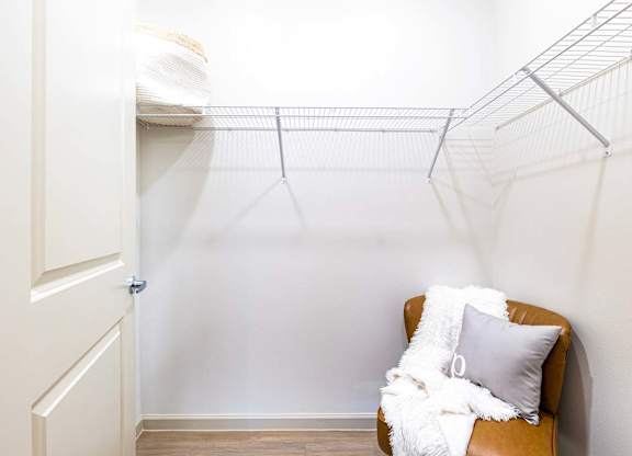 A white chair with a grey pillow and a white blanket is in front of a white wall with a metal shelf above it.