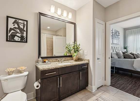 A bathroom with a white toilet, a large mirror, and a bedroom in the background.