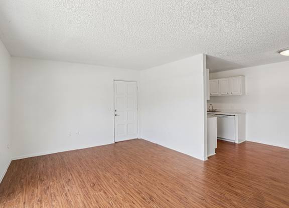 A room with wooden floors and white walls, with a kitchenette visible in the background.