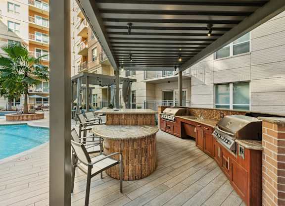 A wooden deck with a table and chairs is surrounded by a pool and apartment buildings.