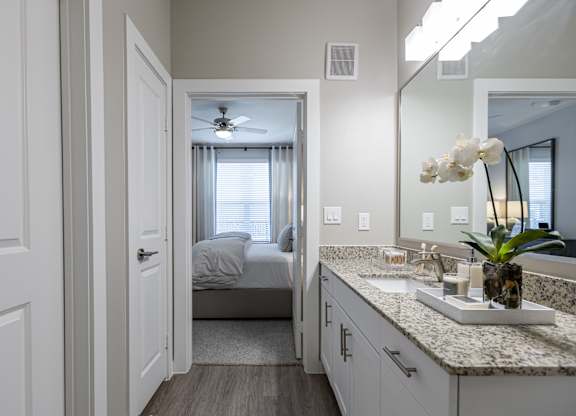 A bathroom with a vanity, mirror, and a bedroom in the background.