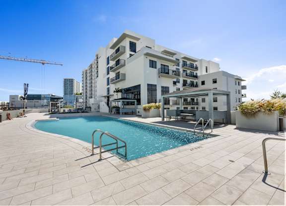 A large swimming pool is surrounded by a tiled patio and a building with balconies.