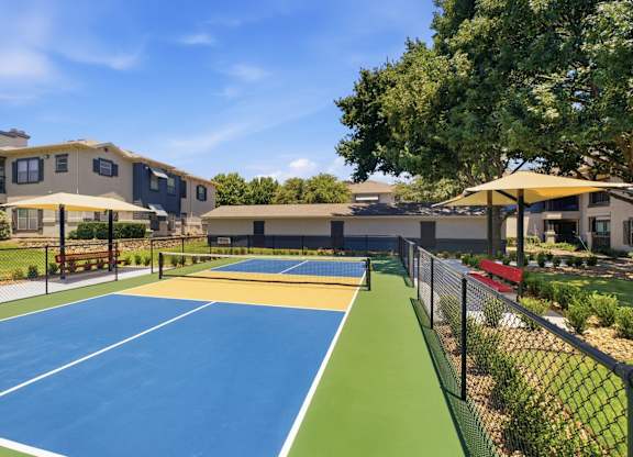 A tennis court is surrounded by a fence and there are two buildings in the background.