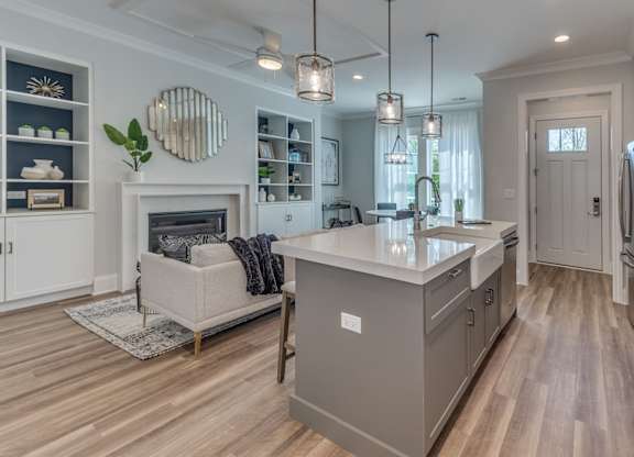 A modern builder grade kitchen with a grey cabinets, wooden floor and front entryway and living room with fireplace in the background.