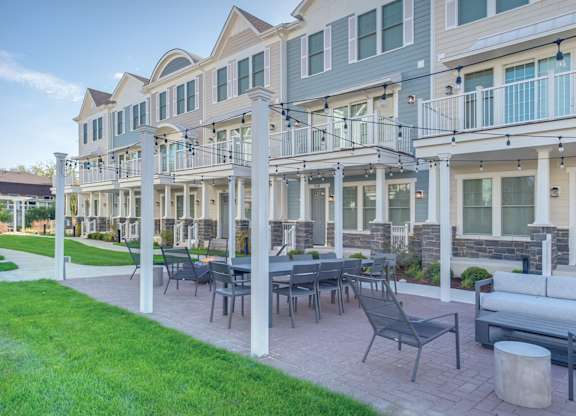 A townhome complex patio with fire pit, table, and chairs on a brick walkway underneath cafe lights.