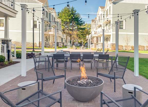 A townhome complex patio with fire pit, table and chairs on a brick walkway underneath cafe lights.