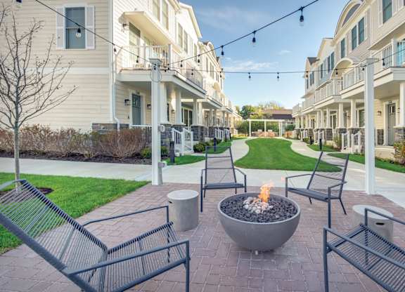A townhome complex patio with fire pit and chairs on a brick walkway underneath cafe lights.