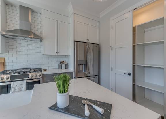 A modern builder grade kitchen with a white countertop and a stainless steel range hood with a double door pantry in the background.