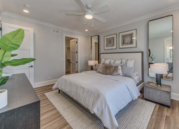 A modern builder grade bedroom with a large bed, a mirror, ceiling fan, and walk in closet.